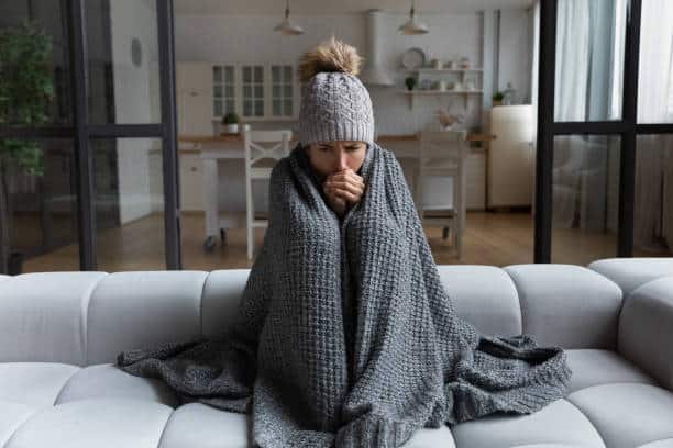 A woman sits on her couch with a knit hat on and a gray blanket covering her up.