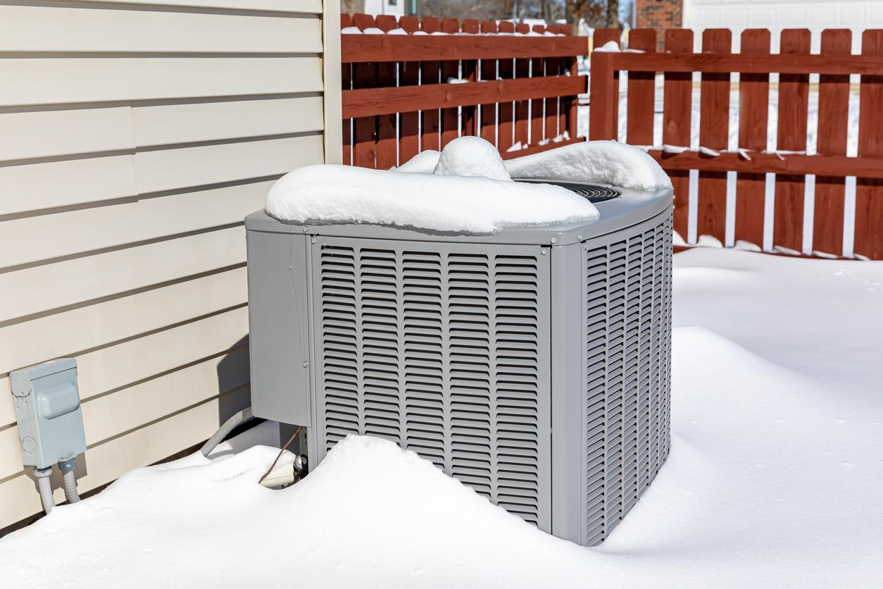 Outdoor AC condenser unit next sits next to home amid snow drifts and a layer of snow on top.