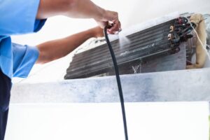 HVAC technician in blue shirt cleans condenser coils on home system